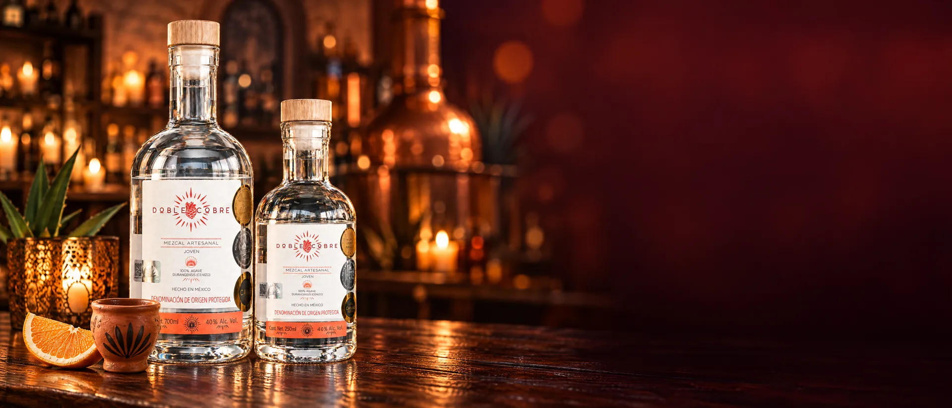 Botellas de Mezcal Doble Cobre de 700 ml y 250 ml en la barra de una mezcalería, junto a una copita de barro y una rodaja de naranja; al fondo, un alambique de cobre