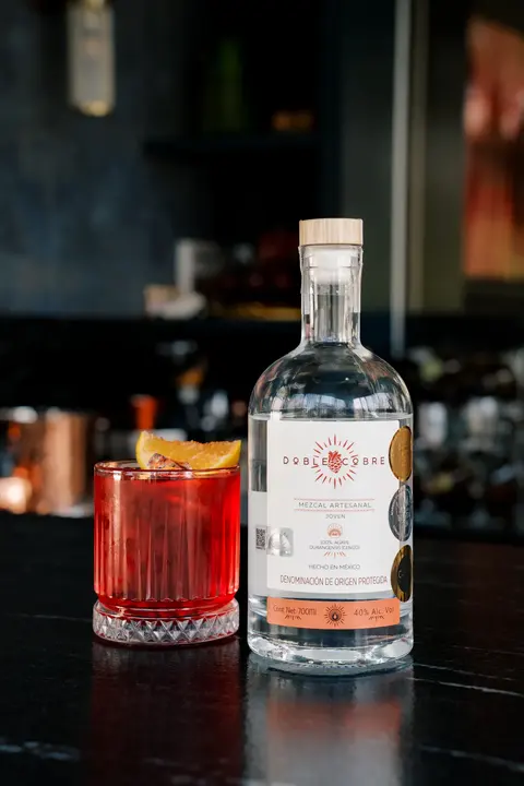 Botella de Mezcal Doble Cobre junto a un coctel rojo en la barra