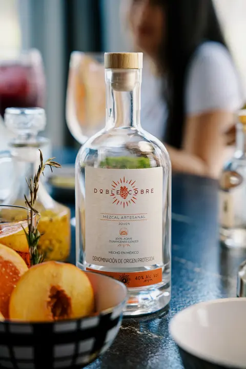 Botella de Mezcal Doble Cobre en una mesa con fruta y copas