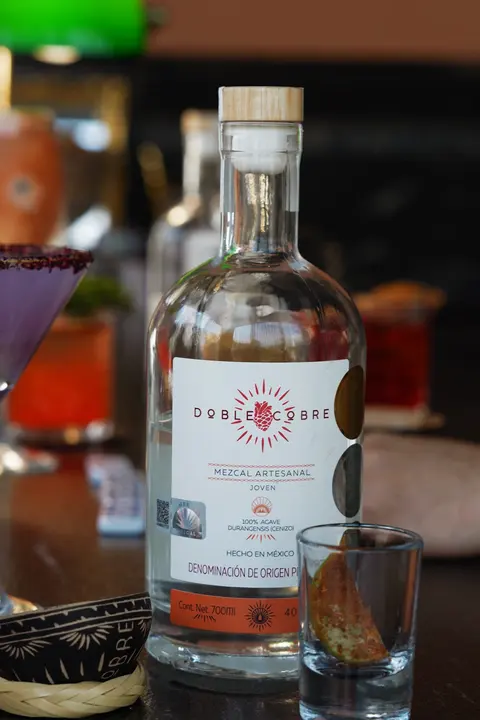 Botella de Mezcal Doble Cobre en una mesa con cocteles y un caballito