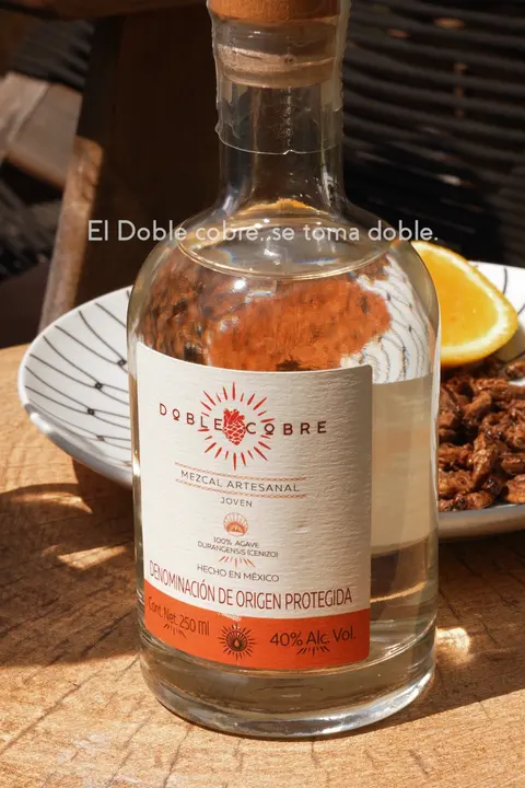 Botella de Mezcal Doble Cobre al sol sobre una mesa de madera con naranja